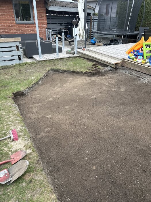 Nygrävd jordplätt bredvid altan; gräsmatta i bakgrunden. Spadar och kvast i förgrunden, redo för anläggning av ny gräsmatta.