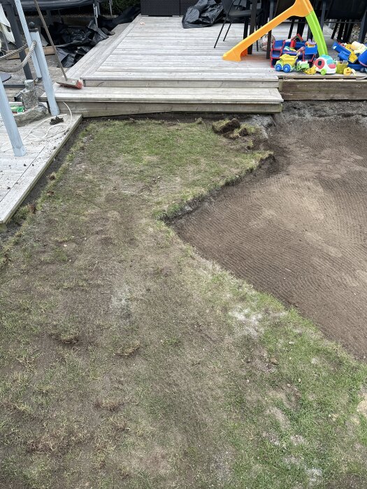 Nyplanterad gräsmatta vid trädäck, synlig jord och leksaker; diskuterar borttagning av gammalt gräs för en kvadratisk form.