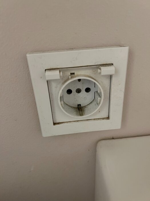 Eluttag i badrumsvägg utan lock, vit ram runt uttaget, delvis smutsig och gammal.