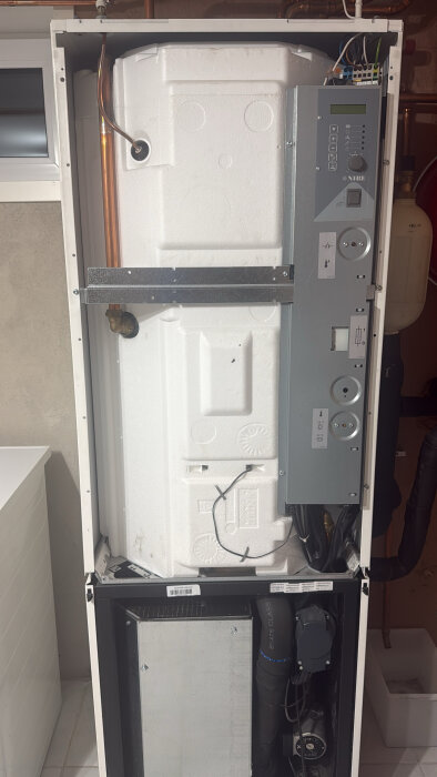 Värmepumpens framsida med styrenhet och röranslutningar synliga, installerad i ett hem.
