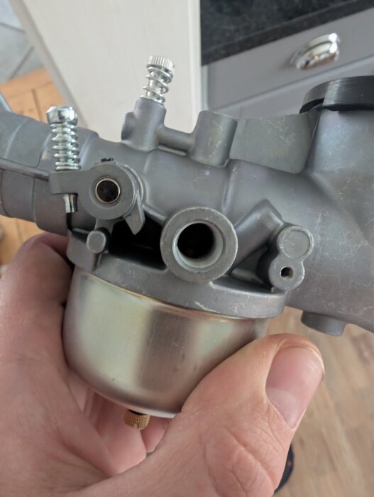 Närbild på en förgasare i metall som hålls i en hand, med synliga skruvar och anslutningar. Bakgrund av trä- och metallmaterial.