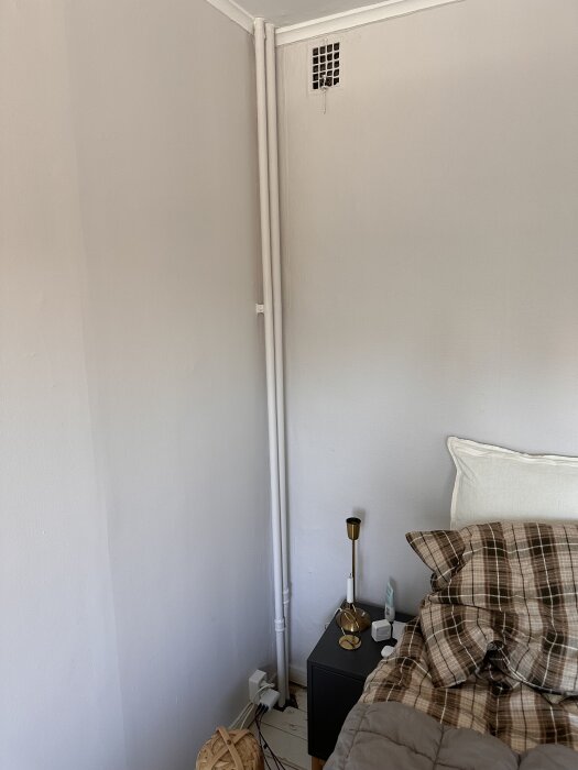Två vita rör i hörnet av ett sovrum, går från golv till tak nära en väggventilation. Säng och nattduksbord synliga i förgrunden.