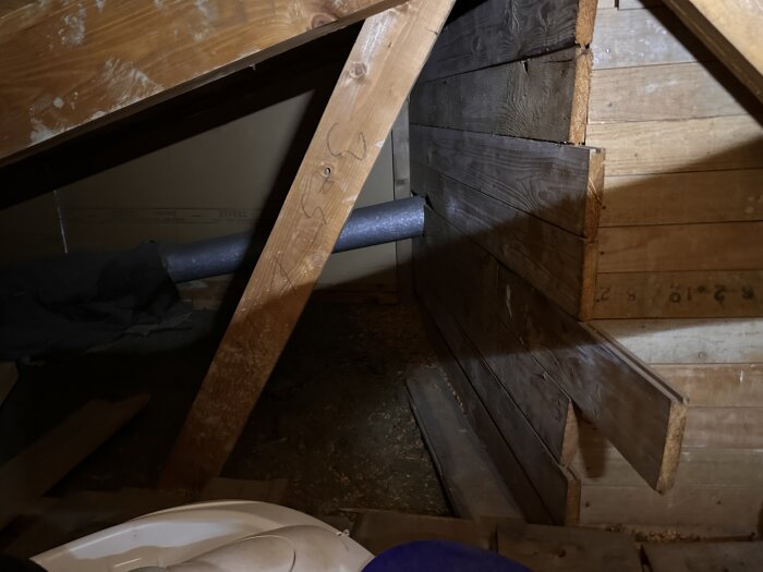 Vind med synliga trästöd och en grå ventilationsrör som löper genom träväggar. Omgiven av olika byggmaterial och låg belysning.