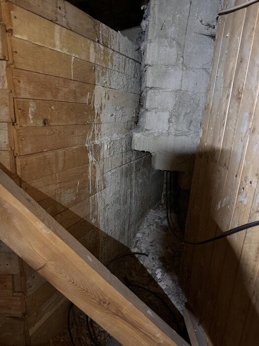 Trångt utrymme med trä- och betongväggar, sprucken puts och kablar som löper längs golvet.