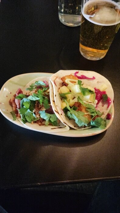 Två tacos på en tallrik, en med halloumi och färska örter, och en med oxbringa, serverade på ett mörkt bord bredvid ett glas öl.