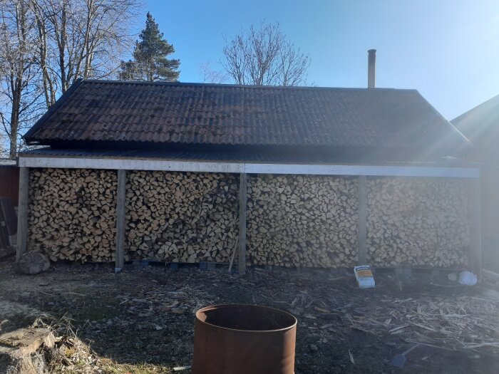 Vedstapel under ett takkonstruktion, utomhus i soligt väder. Marken framför har träflisor och en rostig tunna syns i förgrunden.