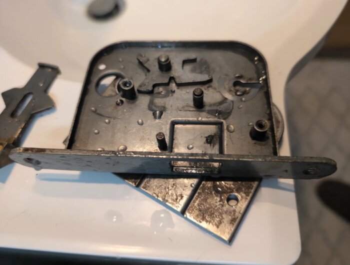 Närbild på ett gammalt låshus av metall, med en slät yta och flera fästhål, placerad på ett vitt underlag.
