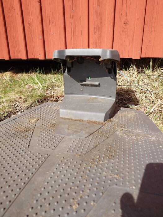 Grå plastdel vid röd träpanel utomhus, omgiven av gräs och jord.