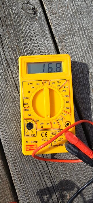 Gult multimeter som visar värdet 16.8 på en träyta, med röda och svarta testsladdar inkopplade.