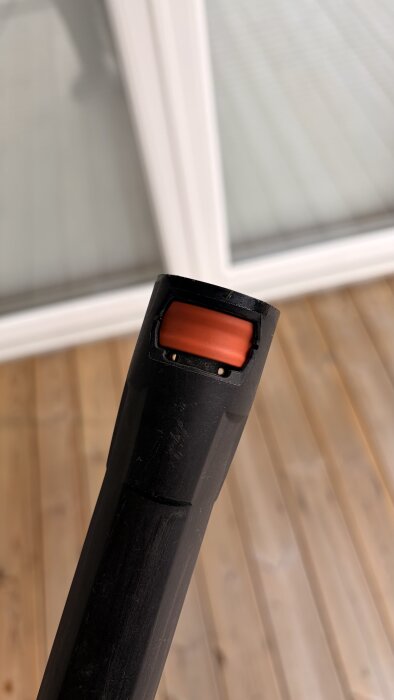 Svart munstycke med orange plastfäste från en högtryckstvätt, fotat mot trätrall och glasdörr i bakgrunden.