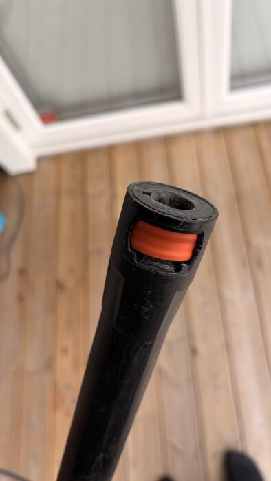 Svart högtryckstvättslang med orange plastfäste och två genomgående hål på änden, liggande på ett trägolv.