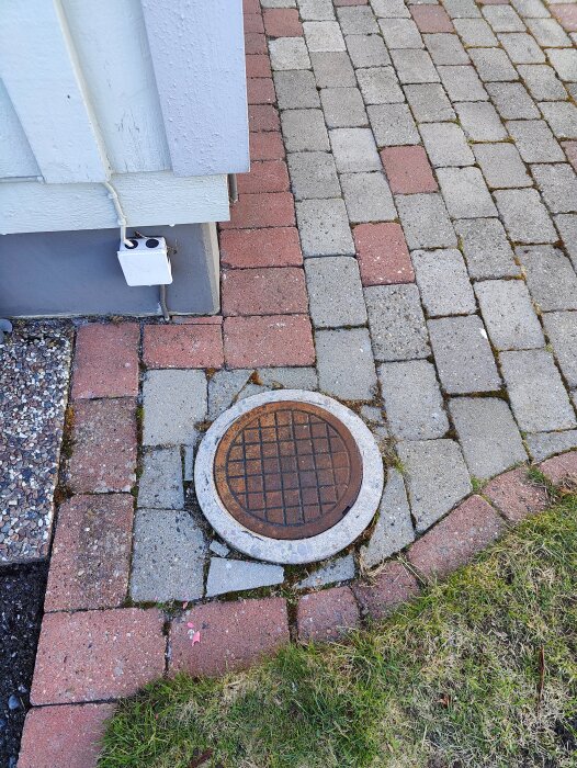 Brunnslock på marken omgiven av marksten i olika färger vid husvägg, del av en trädgård med gräsmatta synlig i hörnet.