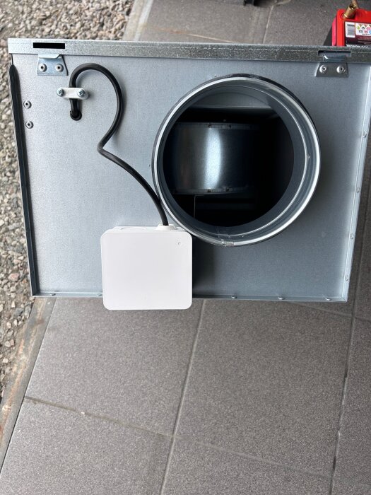 Ventilationsfläkt i metall, med synlig kabel och montering, placerad på grå plattor.