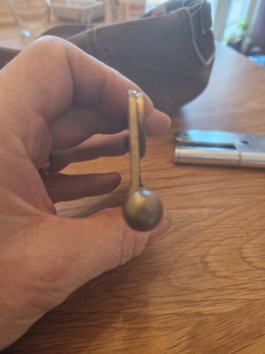 Närbild på ett dörrgångjärn från sidan, håller det mellan fingrarna på ett träbord. Andra verktyg ses suddigt i bakgrunden.