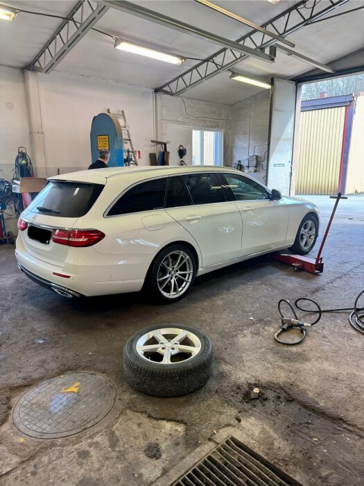 Vit station wagon i verkstad med sommardäck på. Lyft och verktyg i bakgrunden. Ett avtagna vinterdäck ligger på golvet.
