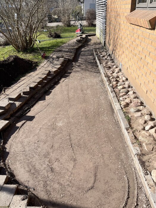 Förberedd gång med grus och kantsten vid husvägg, verktyg i bakgrunden. Fråga om stenläggningens startpunkt och minimering av kapning.