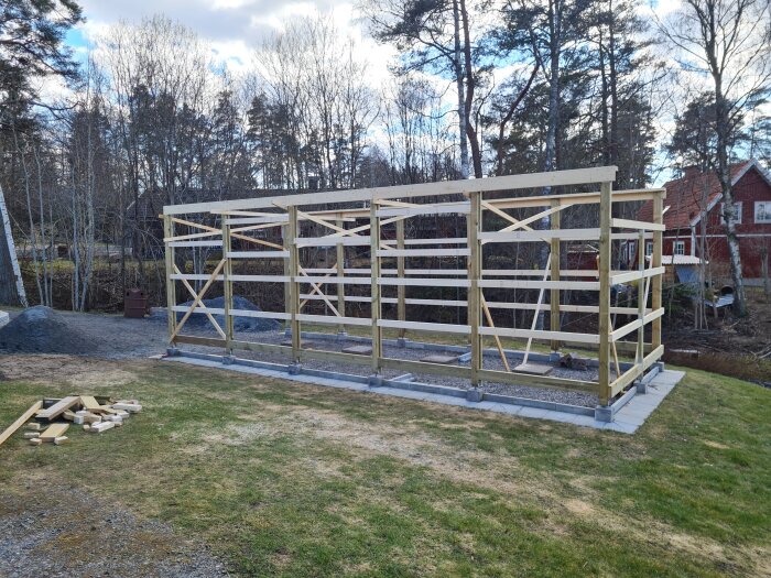 Träram med reglar för ny byggnad, bärlina och hammarband installerade, omgiven av gräsmatta och träd, förberedelser för montering av takstolar.