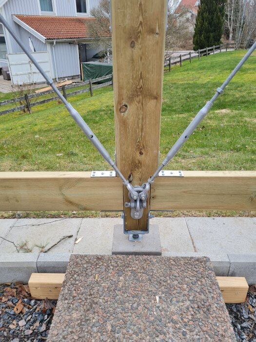 Trästomme med fastsatta vantskruvar och gängade stänger, del av ett byggprojekt. Hus och grön gräsmatta syns i bakgrunden.