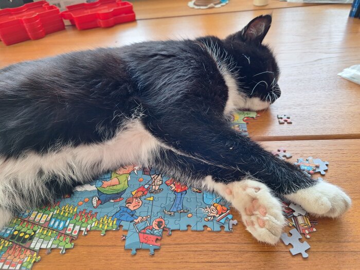 En svartvit katt sover på ett nylagt pussel på ett träbord.