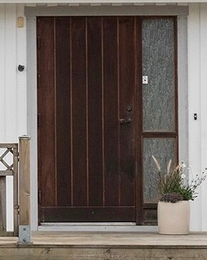 Trädörr med sidofönster av frostat glas, placerad på en veranda med vit fasad. Kruka med växter står bredvid dörren.