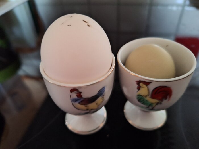 Ägg i äggkoppar med tuppmotiv, ett stort och ett pyttelitet, på köksbänk.