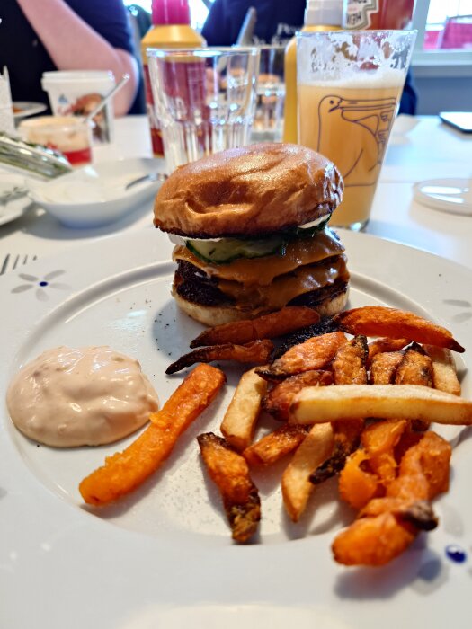 Hamburgare med ost och gurka på en tallrik tillsammans med sötpotatispommes och vanlig pommes, samt en klick sås. Drycker syns i bakgrunden.