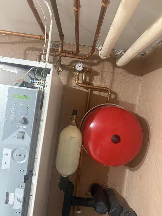 Värmepump med kopparrör, tryckmätare och röd expansionstank på vägg i tekniskt rum.