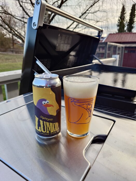 En ölburk och ett glas med öl står på ett stekbord utomhus, omgivet av vinterklädda träd och byggnader i bakgrunden.