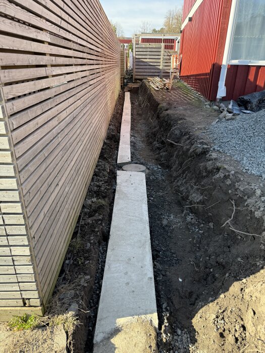 Schaktad mark med långsmal betongplatta och synlig betongbrunn, intill ett rött hus och trästaket. Förberedelse för ny stödmur.