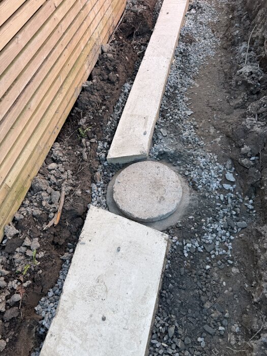 Betongbrunn omgiven av grus och jord på plats där en ny stödmur ska byggas; sidor av trä och betong syns. Brunnen har en diameter på 30 cm.