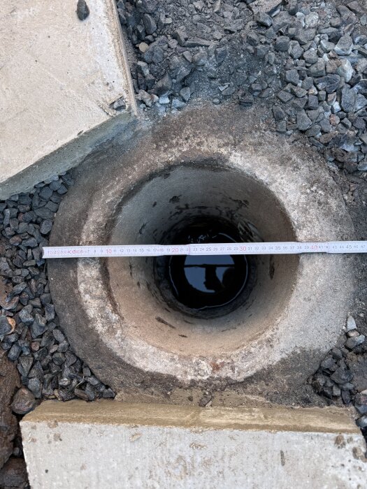 Betongbrunn med innerdiameter på 30 cm omgiven av grus och betongplattor, ett måttband är placerat över öppningen.