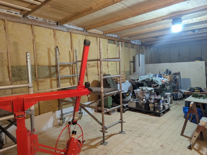 Verkstad under renovering med isolerade väggar, byggställningar, verktyg och material på pallar, fokus på en röd lyftanordning i förgrunden.