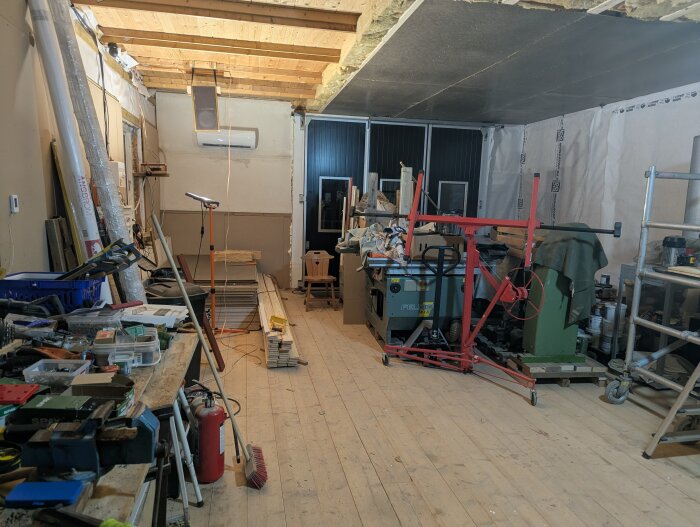 Verkstad under konstruktion med träbjälklag, verktyg, maskiner och byggmaterial på golv och pallar. Kaosartad arbetsyta med verktygsbänkar.