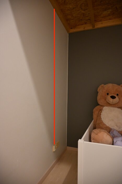 Vägg med orange linje från ett eluttag upp till taket, teddybjörn bredvid vit möbel, märkt för fräsning av flexslangsspår.