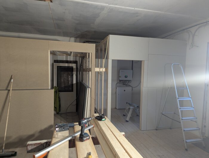 Byggarbetsplats med väggkonstruktion och verktyg, inklusive en stege och en borrmaskin, i en verkstad under konstruktion.
