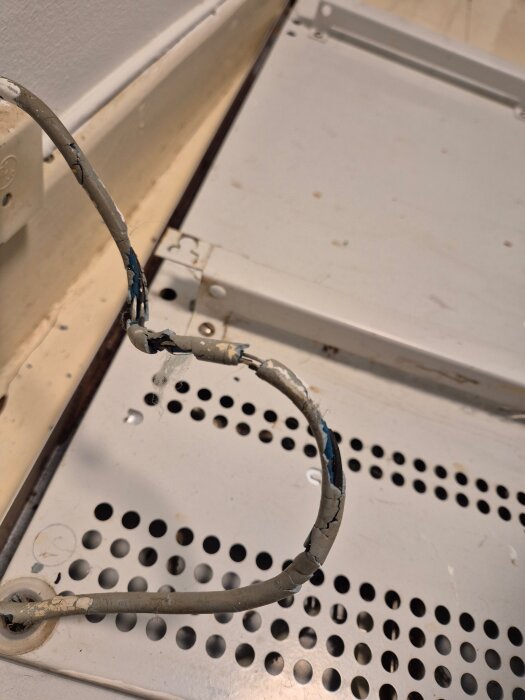 Sliten elektrisk kabel med skadad isolering, borttagen från en gammal elradiator, ligger på ett perforerat metallgolv.