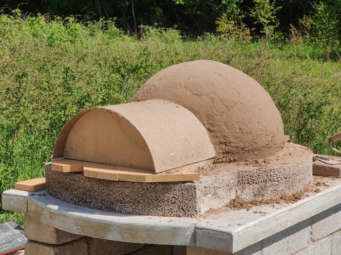 Halvfärdig vedeldad utomhusugn med kupolform, byggd på en bas av sten och sand, omgiven av grönska i bakgrunden.