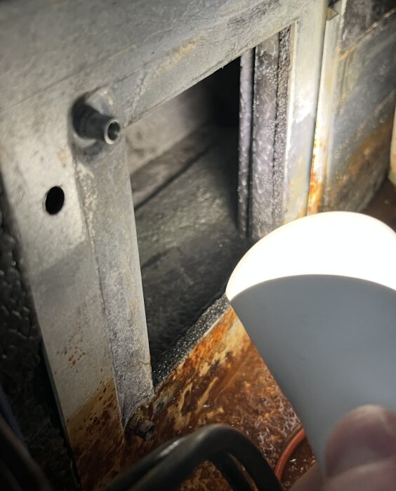 En ficklampa lyser in i en rostig metallöppning fylld med smuts i en form av panel.