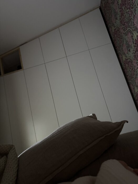 Vita garderobsstommar med frontar monterade, bredvid en blommig tapet, sedda från en soffa med kudde i förgrunden.
