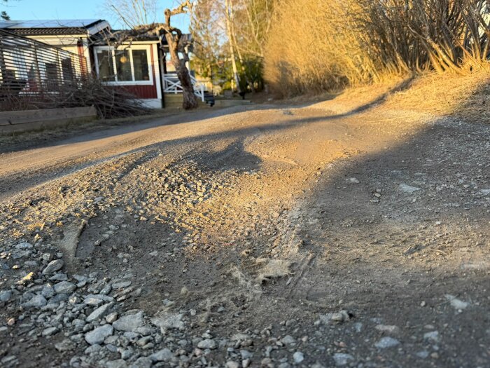 Grusad uppfart i behov av reparation, med ojämnheter och slitage. Hus och buskar i bakgrunden i soligt väder.