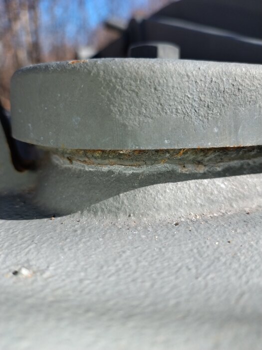 Närbild av en robust, grålackerad metallfäste med viss ytrost, möjlig del av en större lucka med värmetålig packning i bakgrunden.