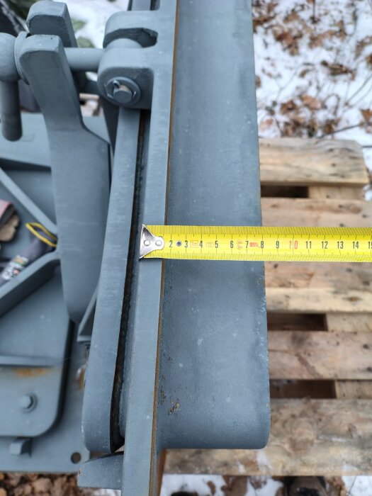 Närbild av en massiv, metalldörr med en snurrhandtag, placerad på en träpall med en måttstock som visar storleken på kanten. Snöig mark i bakgrunden.
