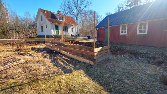 Odlingslåda under konstruktion i en trädgård med trästängsel runt, omgiven av ett rött och ett vitt hus.