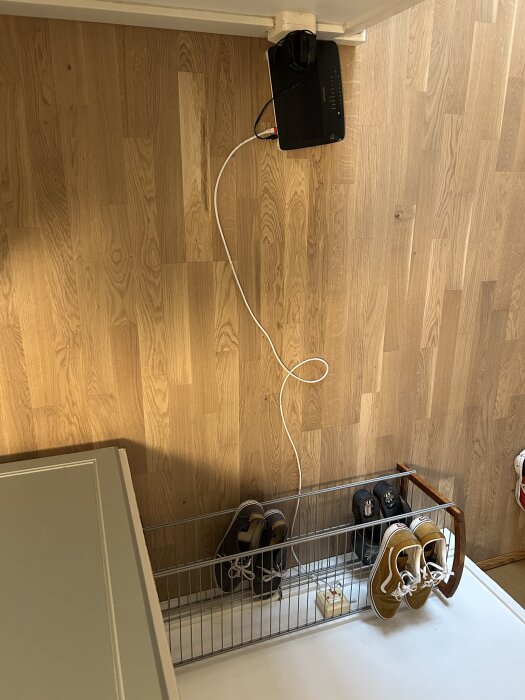 Internetmodem placerat ovanför skohylla med nätverkskabel exponerad längs väggen, nära träningsskor och ett eluttag.