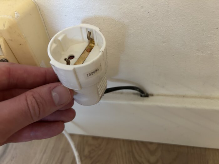 En hand håller i ett trasigt eluttag med synliga mässingskontakter, mot en vit vägg.
