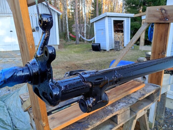 Nymålad axeldel upphängd i ställning utomhus, lackad med svart 2K-färg. Bakom syns verktygsskjul och vedhög.