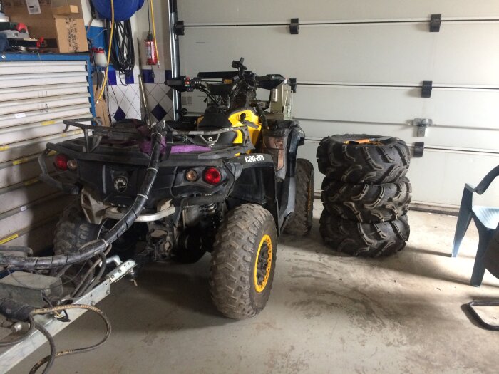 ATV parkerad i ett garage med en trailer kopplad och en stapel av grovmönstrade däck bredvid.