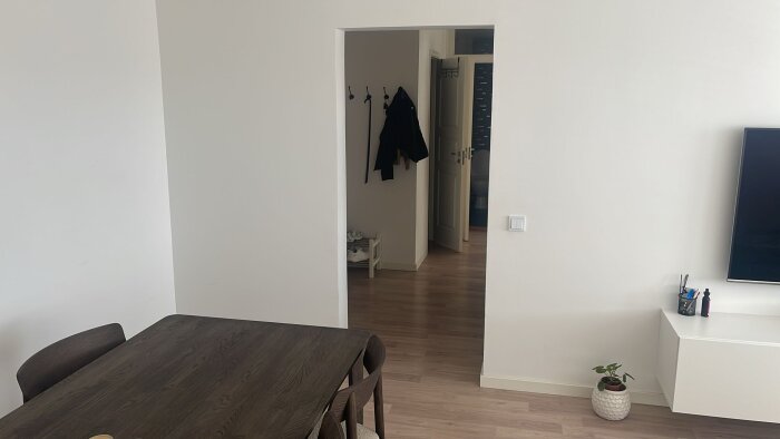 Öppning i vit betongvägg mellan matsal och hall med jackor hängande, mörkt träbord och svart TV-skärm på väggen till höger.