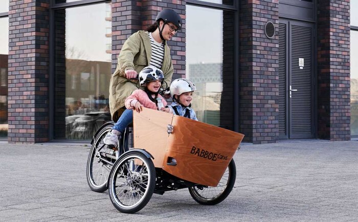 Cykel av modellen Babboe Carve med två barn i lådan, cyklas av en vuxen person, alla bär hjälmar.