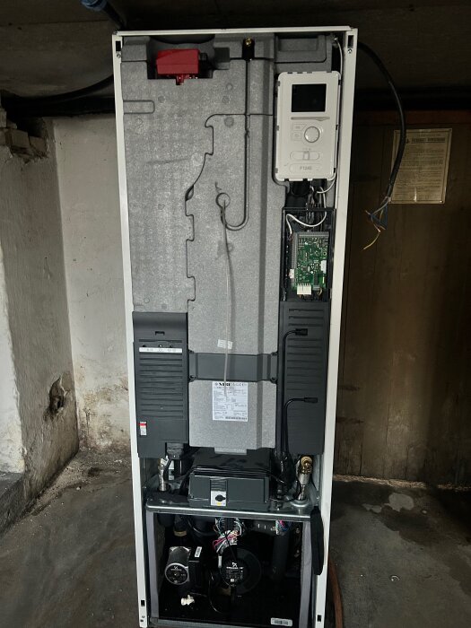 Nedmonterad NIBE F1245-12CU jordvärmepump i källarmiljö, visar pumpens insida med panel och komponenter.